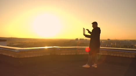 Hacker-using-laptop-on-rooftop-with-city-view-and-forex-chart.-Hacking-and-stats-concept.-A-man-at-sunset-in-slow-motion-writing-software-code-on-a-laptop